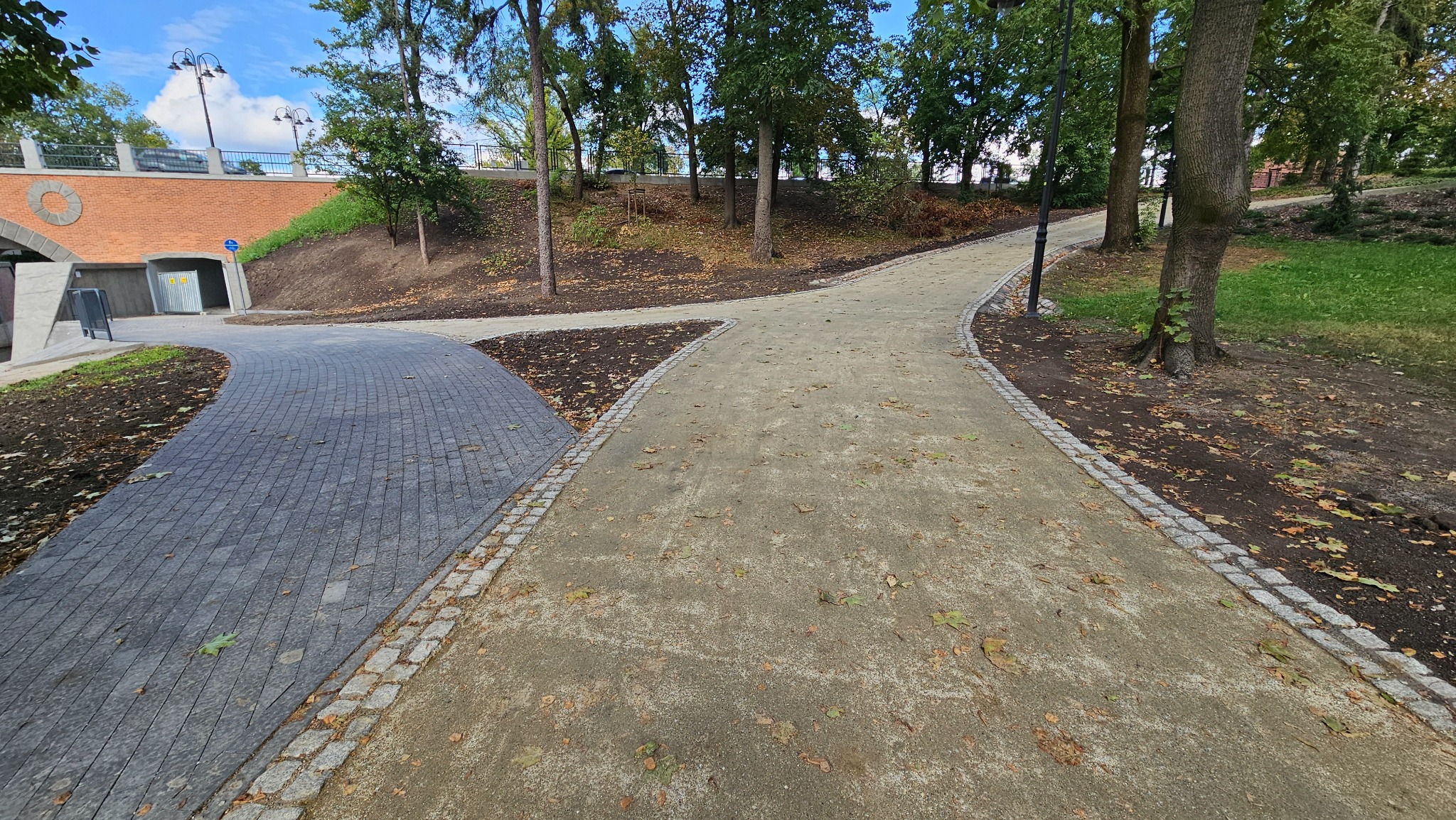 Rozwidlenie ścieżek w parku: z lewej szara kostka brukowa w łuku, z prawej utwardzona droga gruntowa z obrzeżem z kostki granitowej, jesienne liście na ziemi, drzewa w tle.