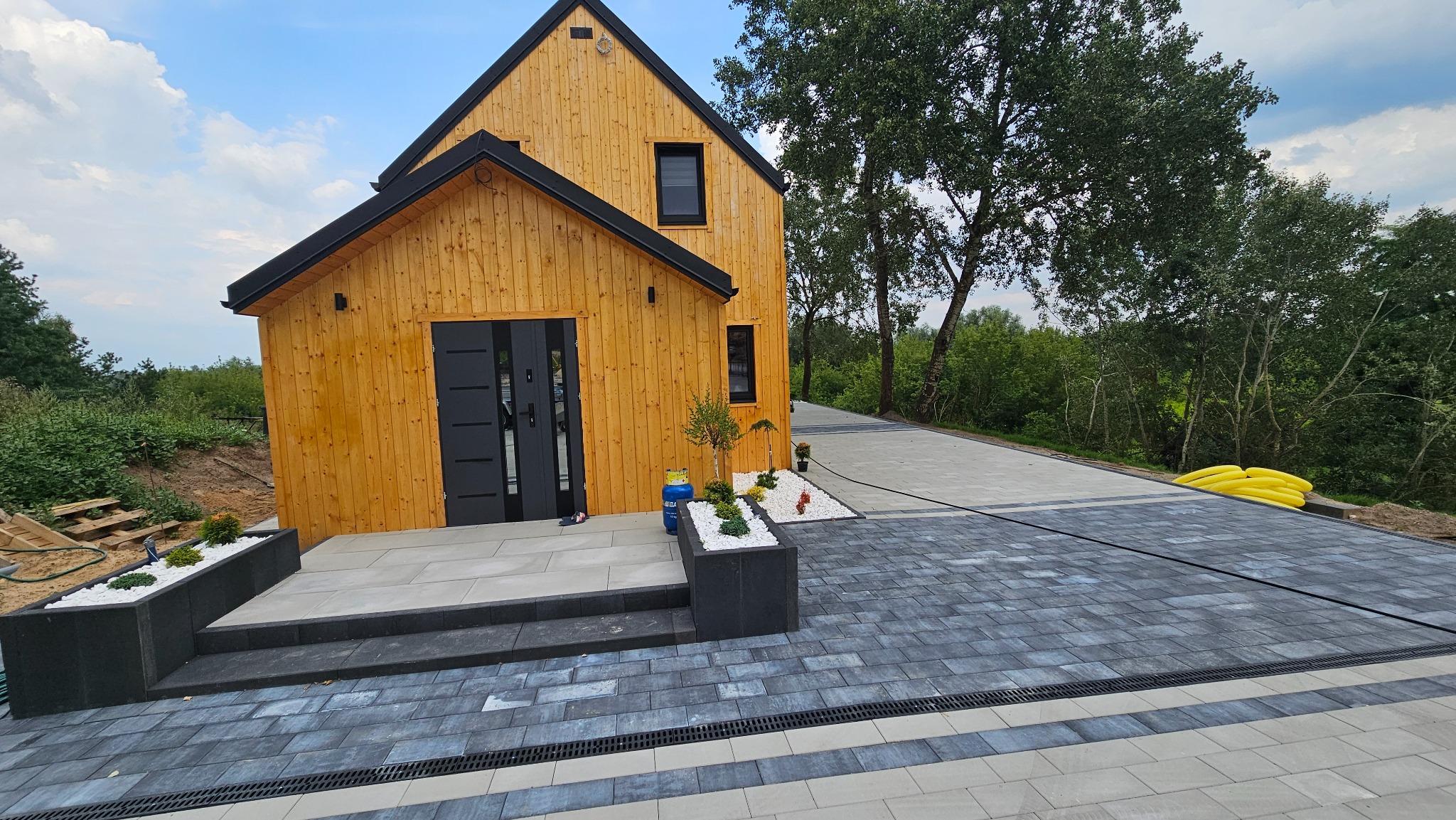 Nowoczesny dom z drewnianą elewacją i ciemnymi oknami, otoczony nowo ułożoną szarą kostką brukową z odpływem liniowym, rabatami z białym kamieniem i zielenią oraz żółtymi rurami w tle.