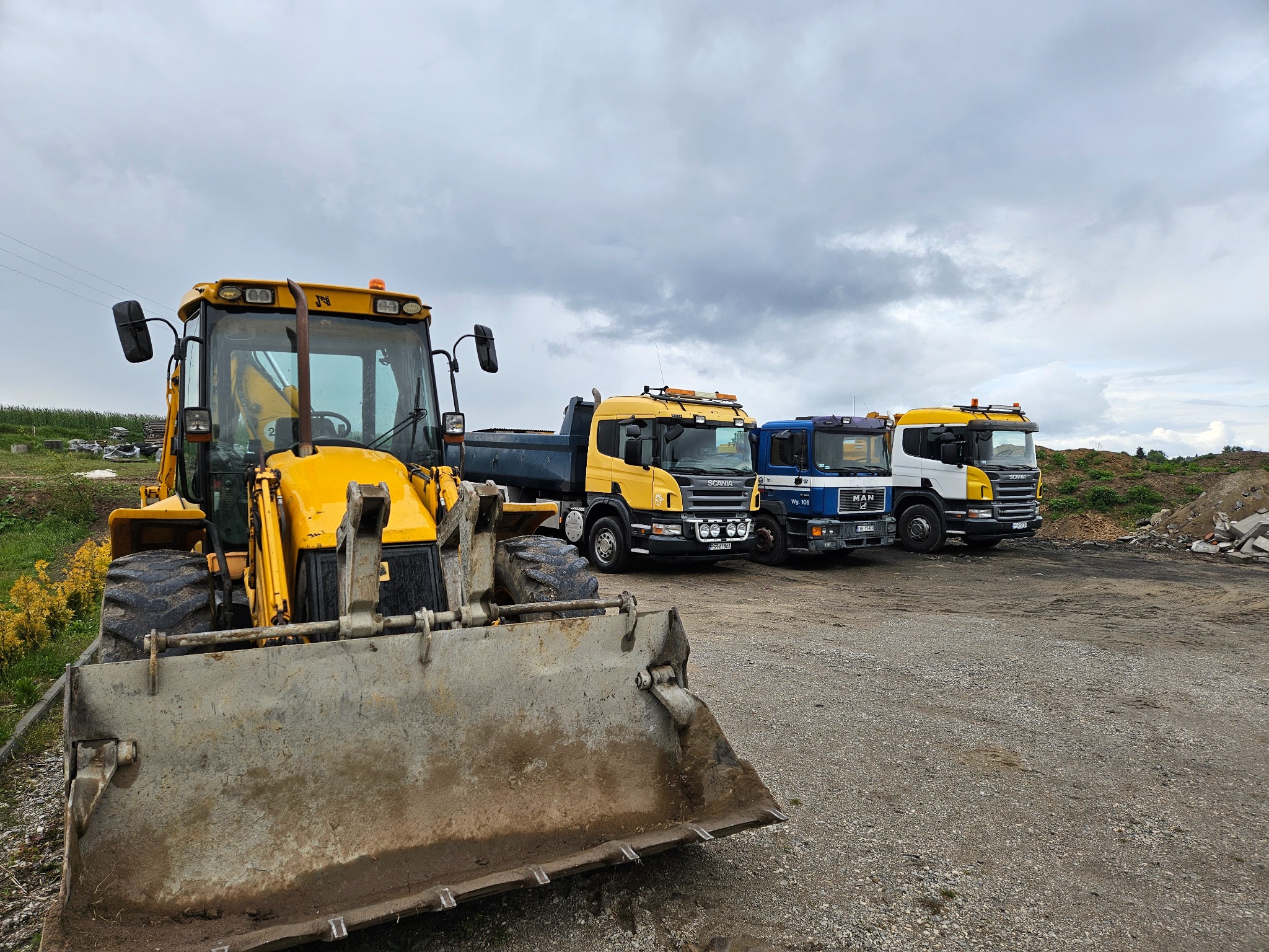 Żółta koparko-ładowarka JCB z brudną łyżką na pierwszym planie, w tle zaparkowane trzy ciężarówki Scania i MAN, pochmurne niebo.