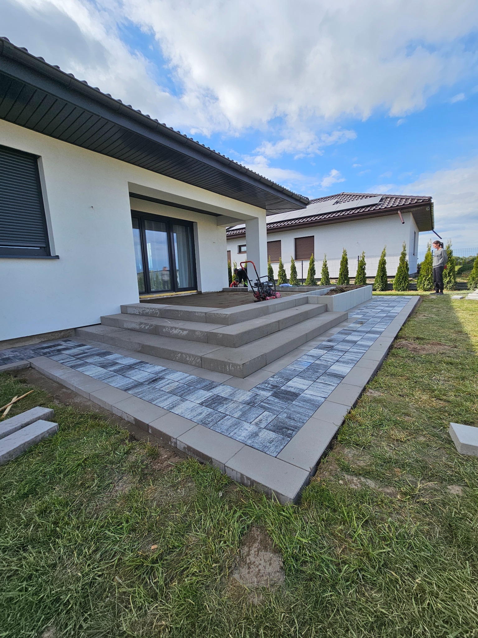 Schody i ścieżka z kostki brukowej prowadząca do domu z białymi ścianami. Częściowo ułożona kostka, na trawniku leżą dodatkowe elementy. Widoczna zagęszczarka.