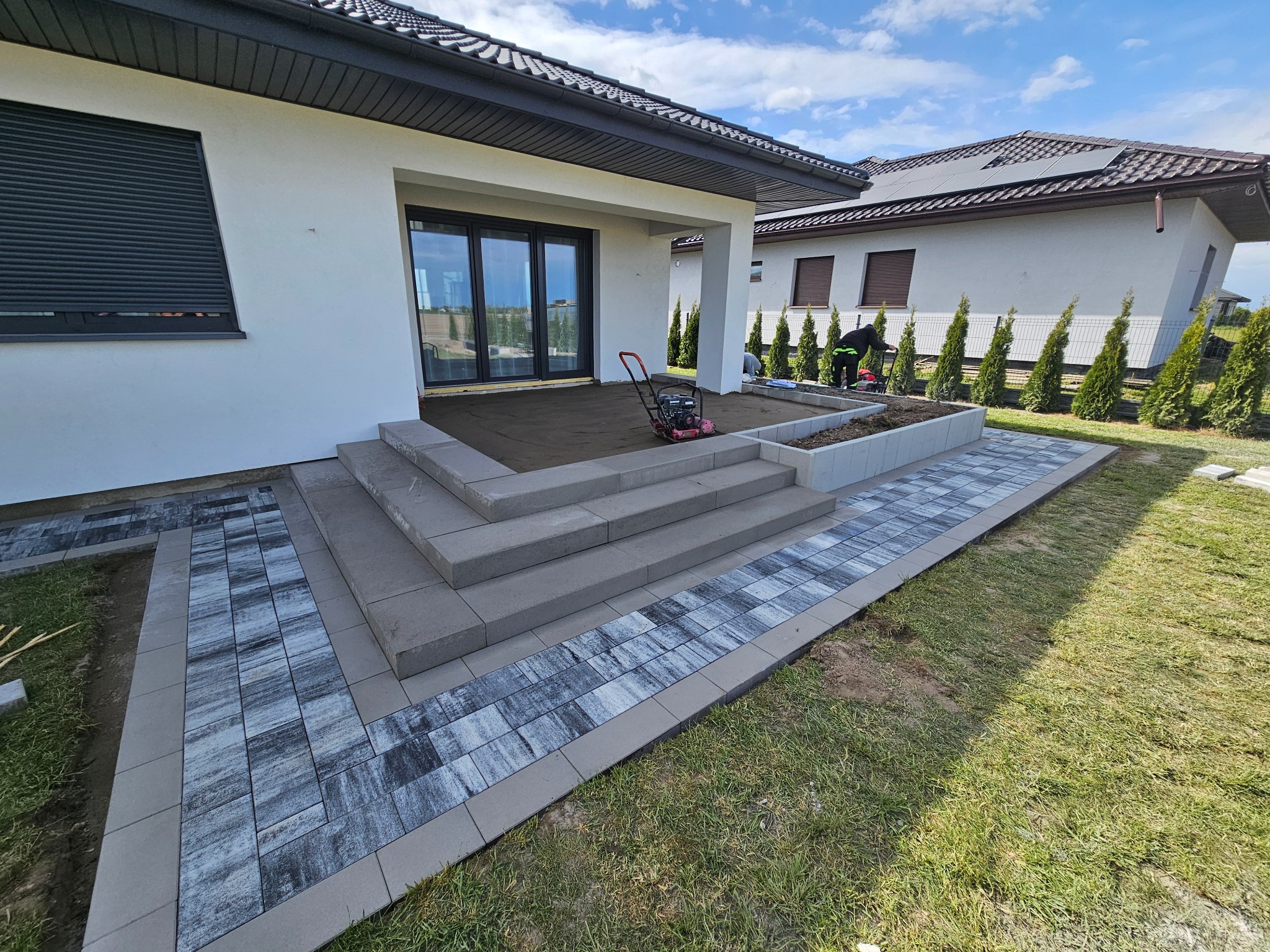 Nowoczesny dom z tarasem w trakcie budowy, widoczne schody z kostki brukowej, przygotowany teren pod taras i pracownik z zagęszczarką.