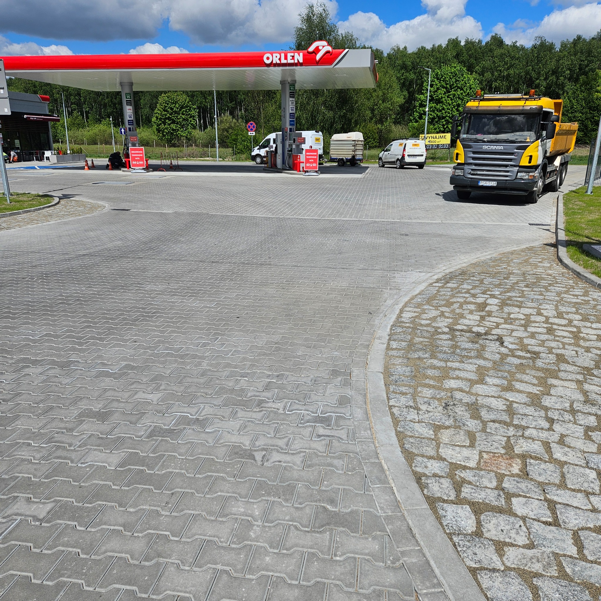 Nowa nawierzchnia z kostki brukowej na stacji paliw Orlen, z żółtą ciężarówką Scania w tle. Widoczna granica między kostką brukową a kamienną.