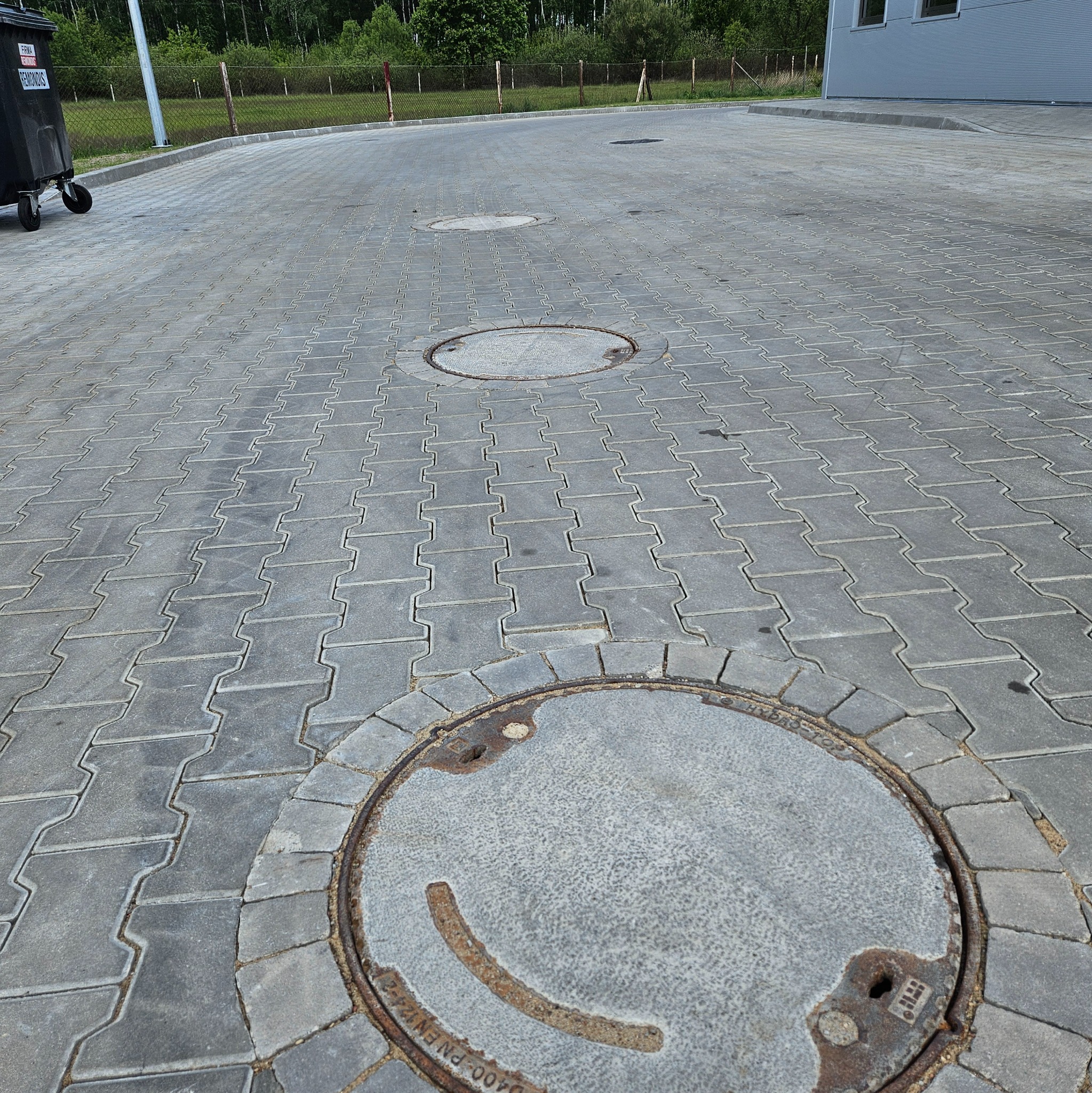 Szara kostka brukowa typu I z trzema włazami kanalizacyjnymi. Widok z góry na utwardzony teren w otoczeniu zieleni i budynku.
