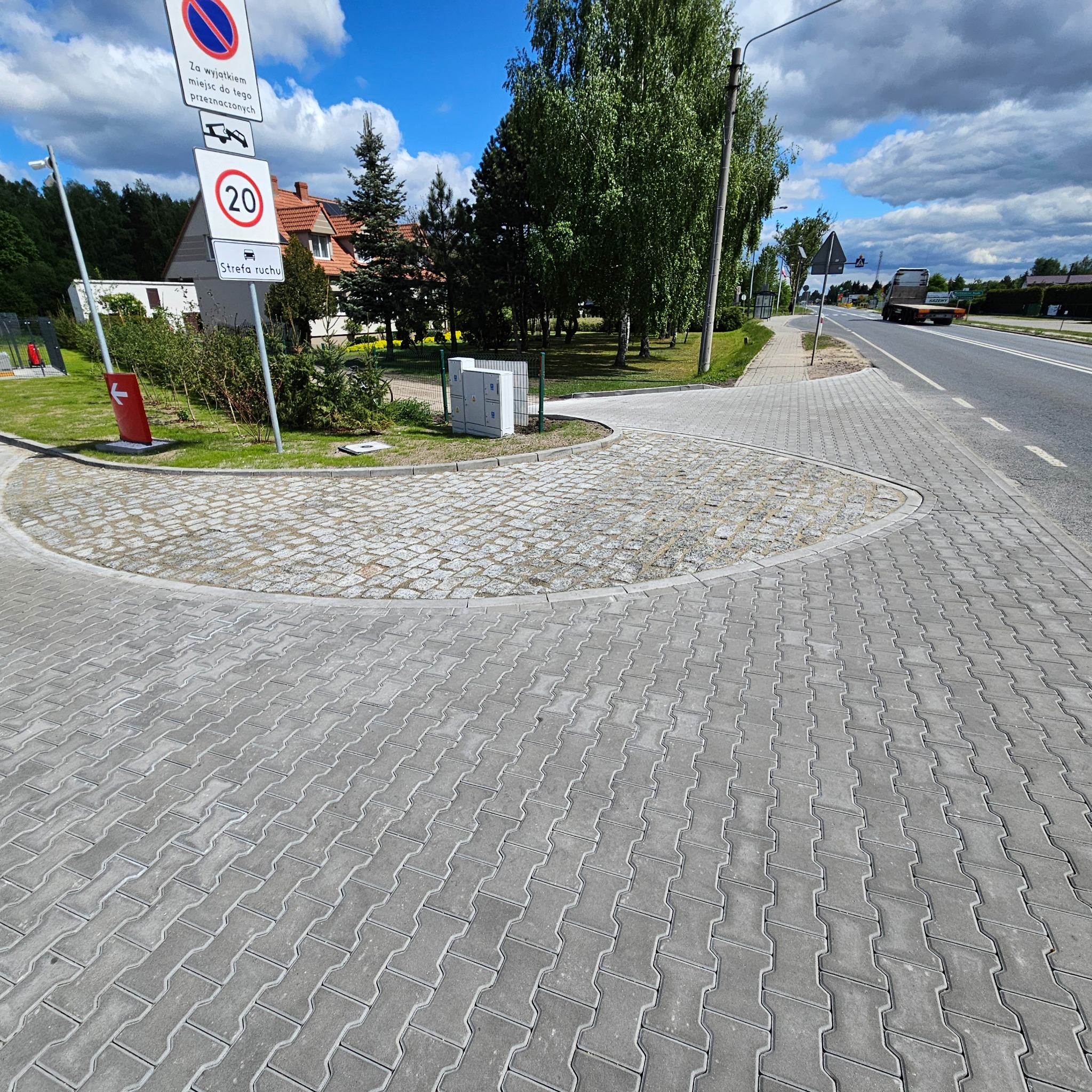 Szara kostka brukowa ułożona na parkingu przy drodze, z widocznymi znakami drogowymi i zielenią. Elementy małej architektury i nowa nawierzchnia.