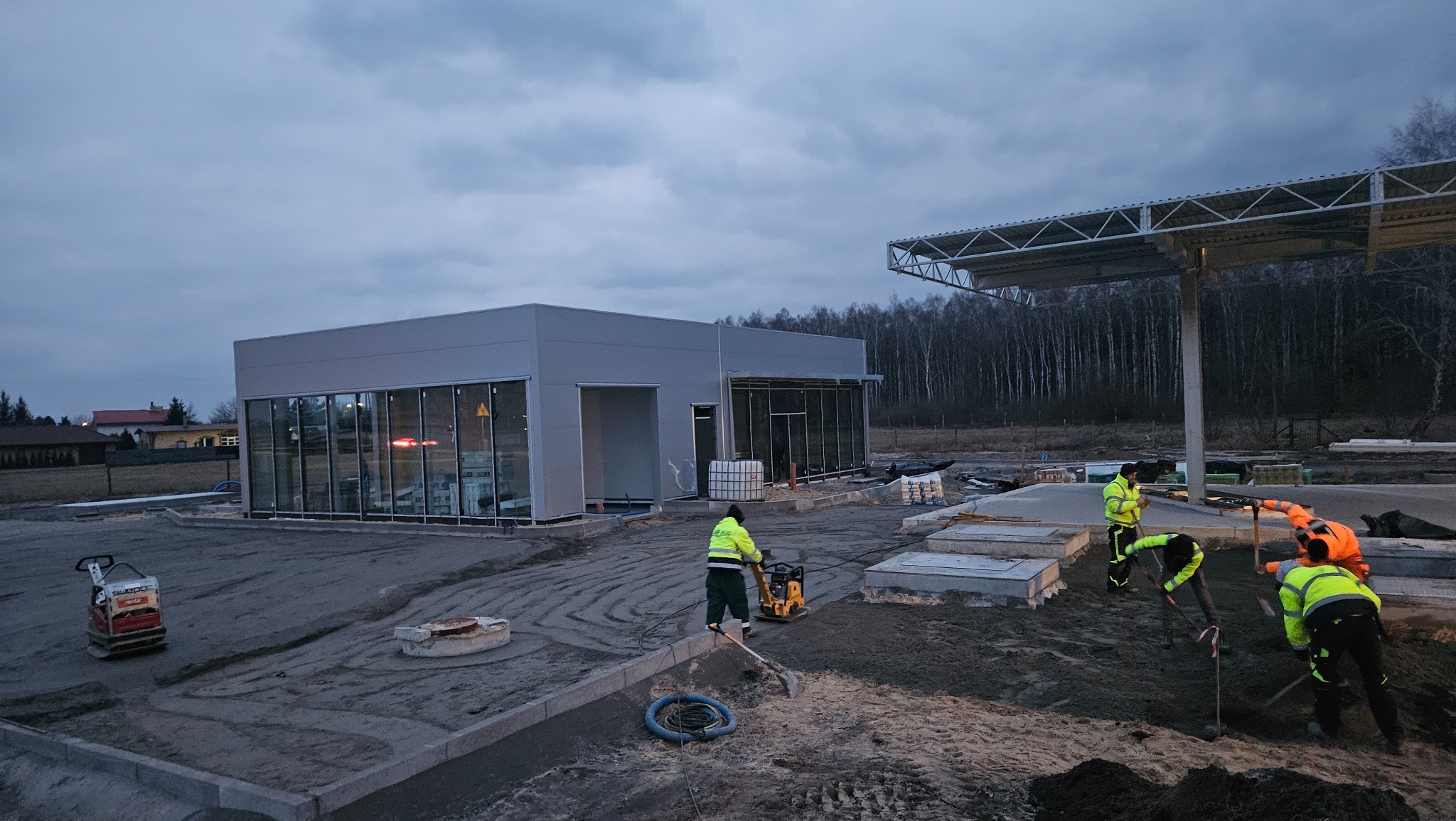 Budowa obiektu usługowego: robotnicy w odblaskowych kamizelkach wyrównują teren podłoża. W tle budynek z dużymi oknami i zadaszenie. Widoczna zagęszczarka.