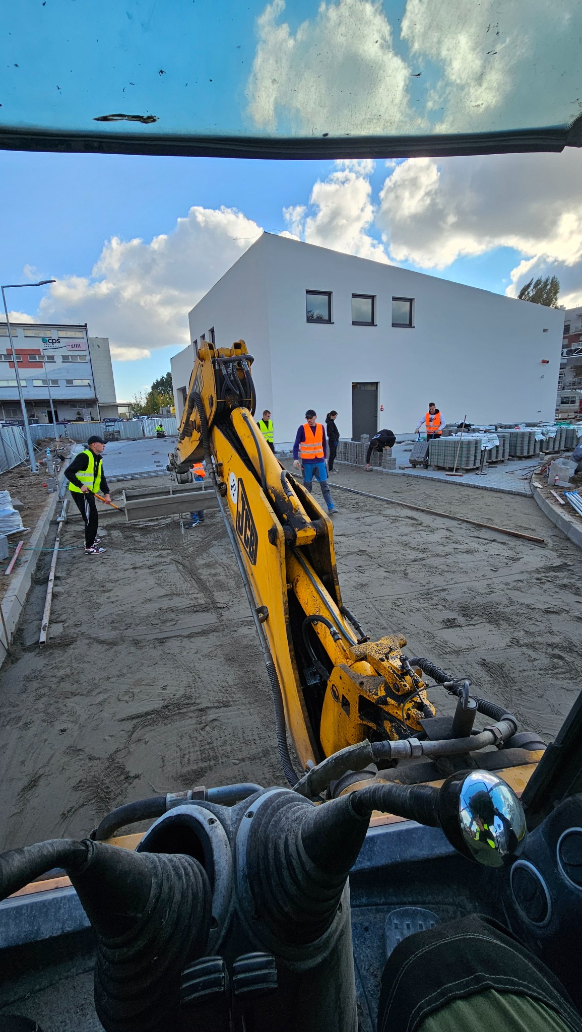 Widok z kabiny koparki JCB na plac budowy: robotnicy w kamizelkach układają kostkę brukową na przygotowanym podłożu, w tle biały budynek i błękitne niebo.