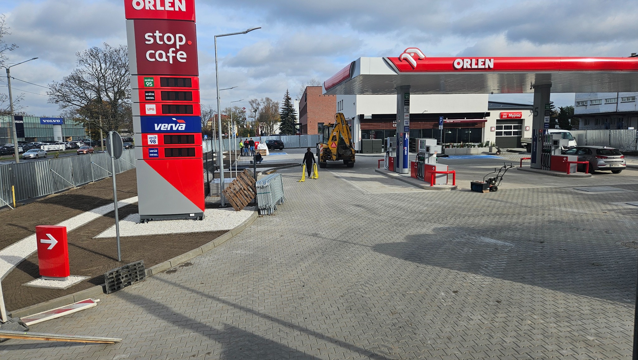 Stacja paliw Orlen po pracach brukarskich. Nowa nawierzchnia z kostki, wykończone rabaty z białym kamieniem. W tle koparka i pracownicy.