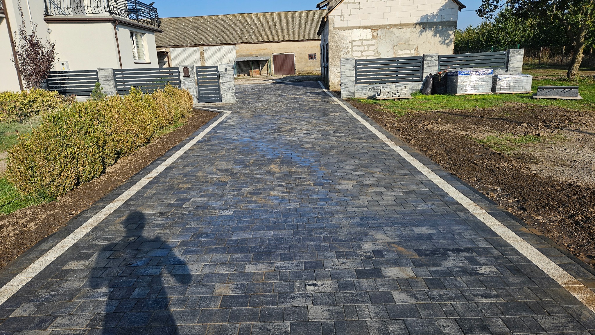 Ułożona, grafitowa kostka brukowa z jasnymi obrzeżami tworzy elegancki podjazd. W tle budynki gospodarcze i nowoczesne ogrodzenie z paletami kostki obok.