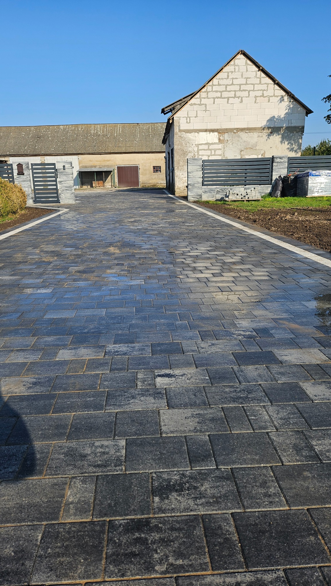 Ułożona, szara kostka brukowa na podjeździe z białymi obrzeżami, prowadząca do zabudowań gospodarczych w słoneczny dzień. Nowoczesne ogrodzenie po bokach.