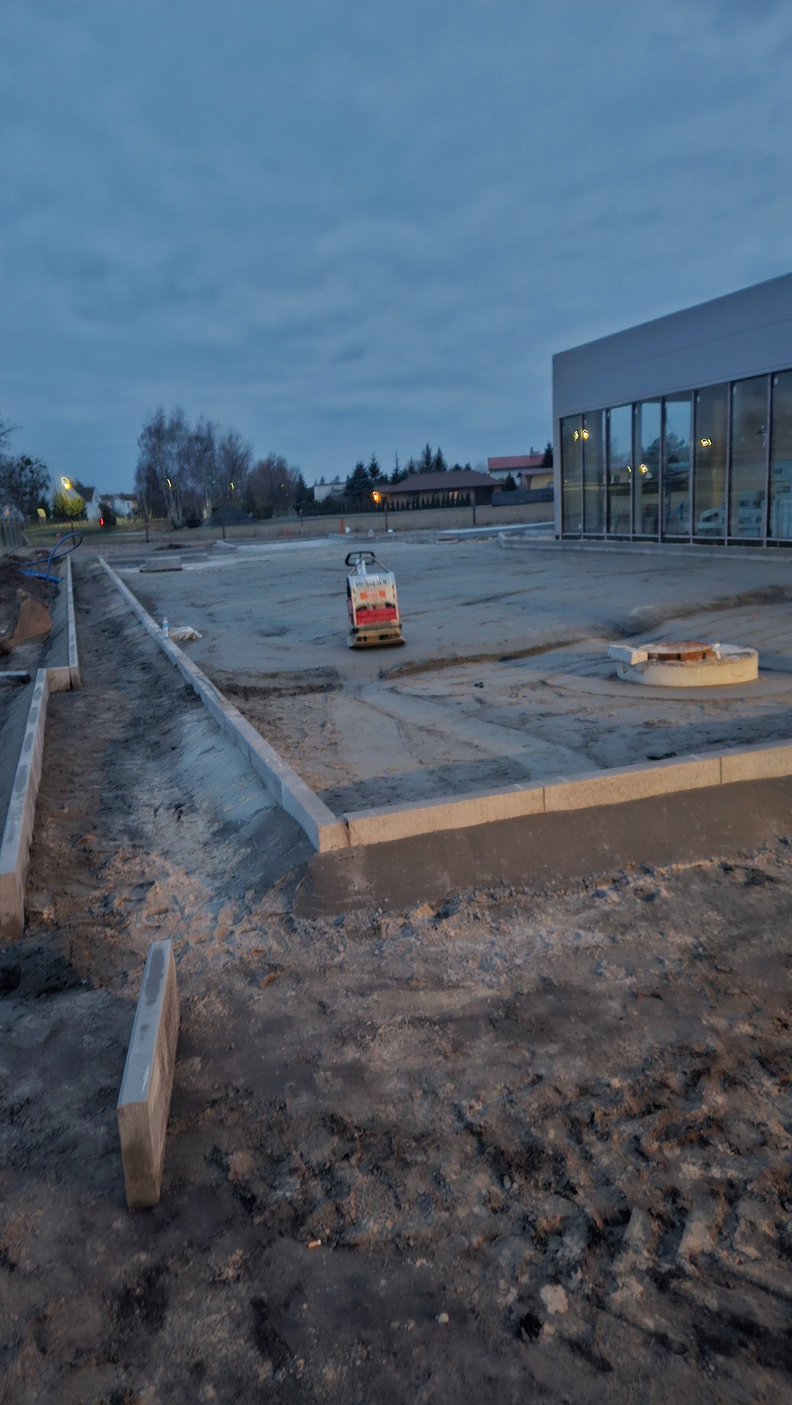 Przygotowanie terenu pod budowę: wyrównany grunt, obrzeża chodnikowe wzdłuż budynku z przeszkleniami, zagęszczarka w tle. Budowa w toku.