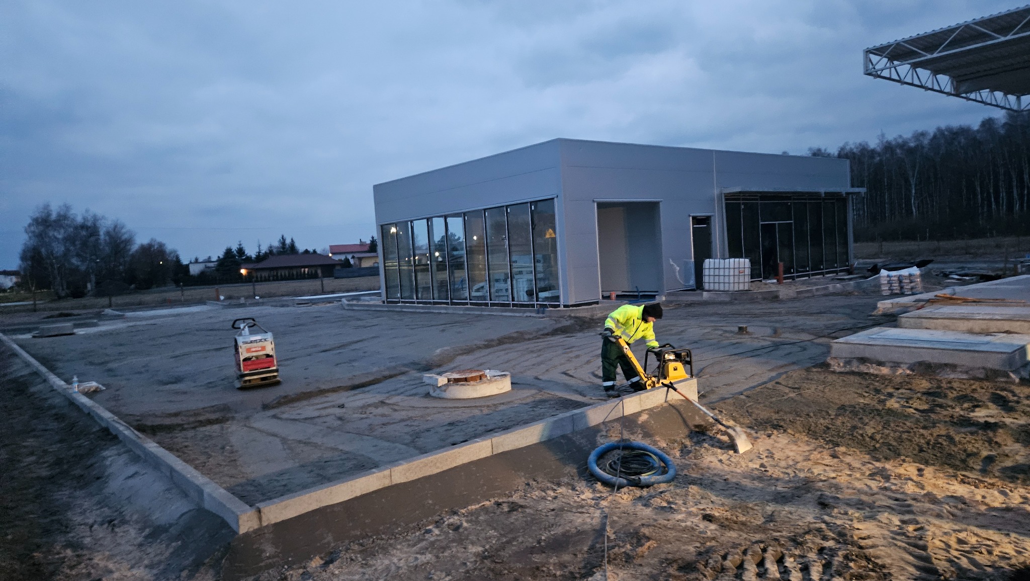 Pracownik w żółtej kurtce zagęszcza piasek zagęszczarką wibracyjną. W tle budynek z dużymi oknami i drugi sprzęt. W tle drzewa.