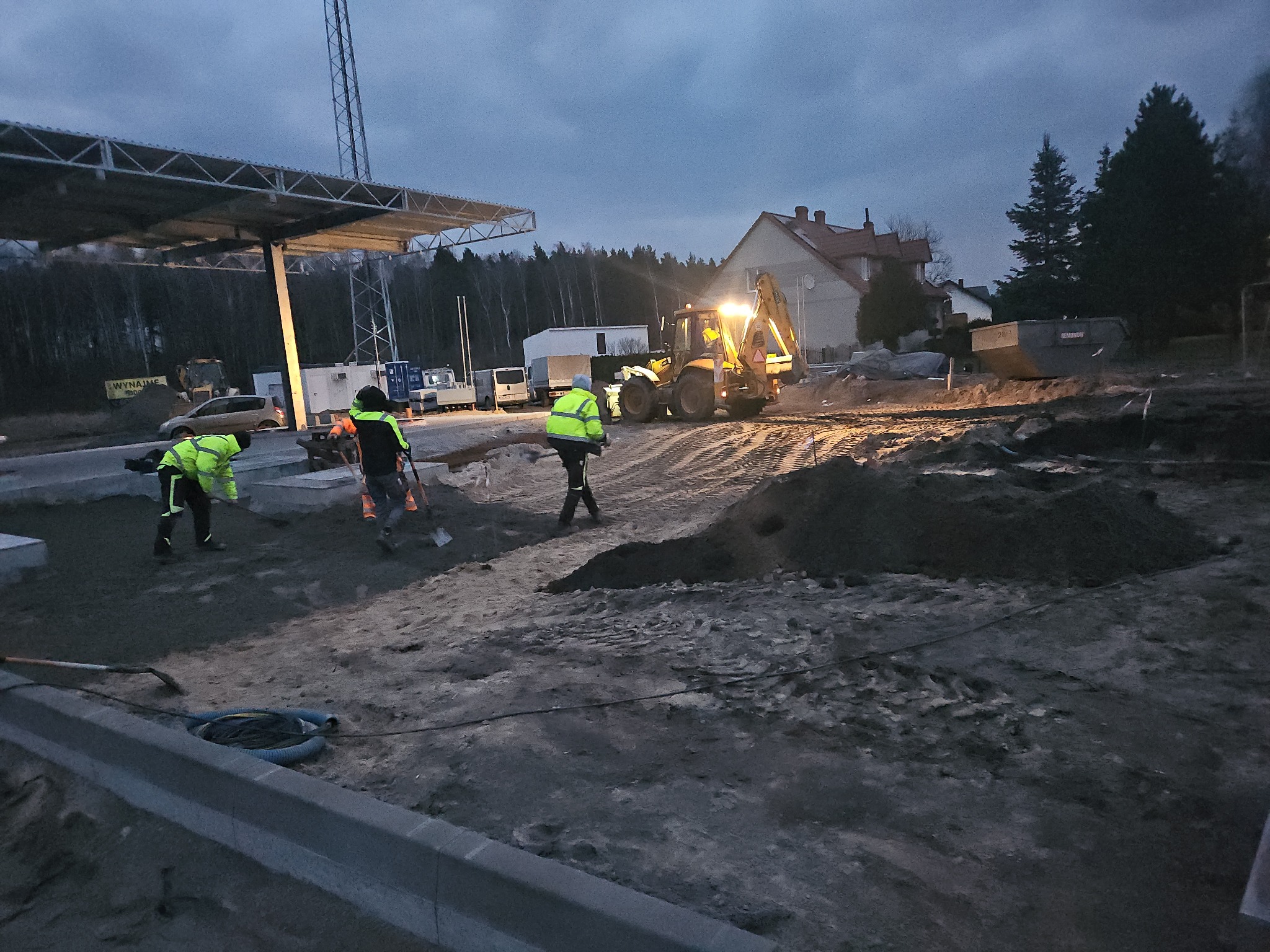 Prace ziemne w Lubaniu: robotnicy w odblaskowych kamizelkach przygotowują teren pod budowę, w tle koparka i budynki. Widoczne krawężniki i hałdy ziemi.
