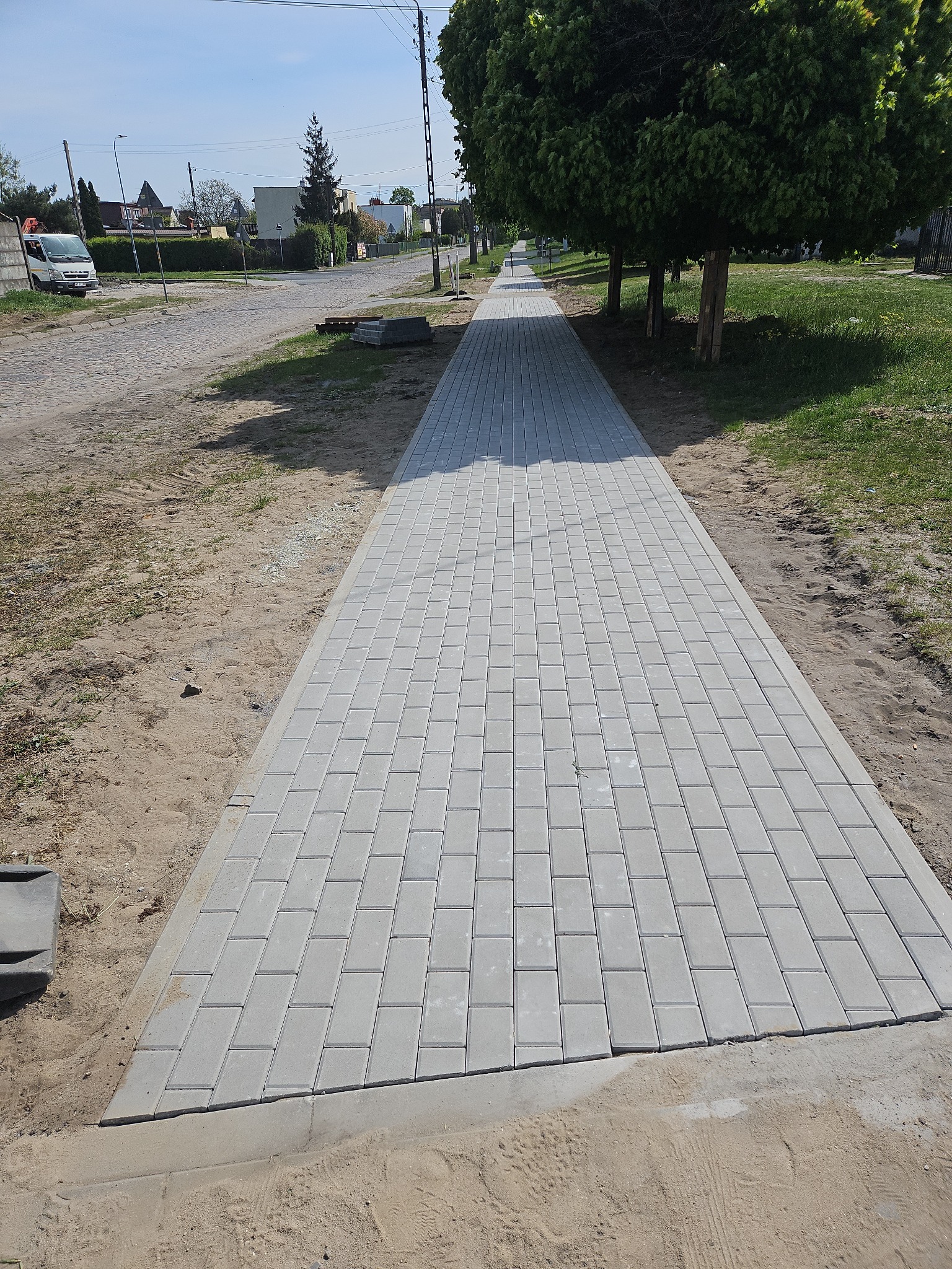 Nowy chodnik z szarej kostki brukowej, świeżo ułożony wzdłuż ulicy z drzewami, z widocznymi pozostałościami piasku po pracach brukarskich. Słoneczny dzień.