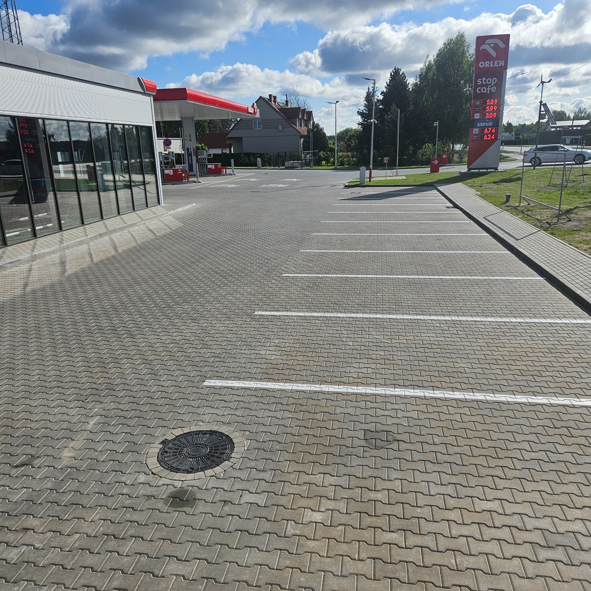 Szara kostka brukowa ułożona na parkingu stacji benzynowej z białymi liniami wyznaczającymi miejsca postojowe. Widoczny właz kanalizacyjny i budynek stacji.