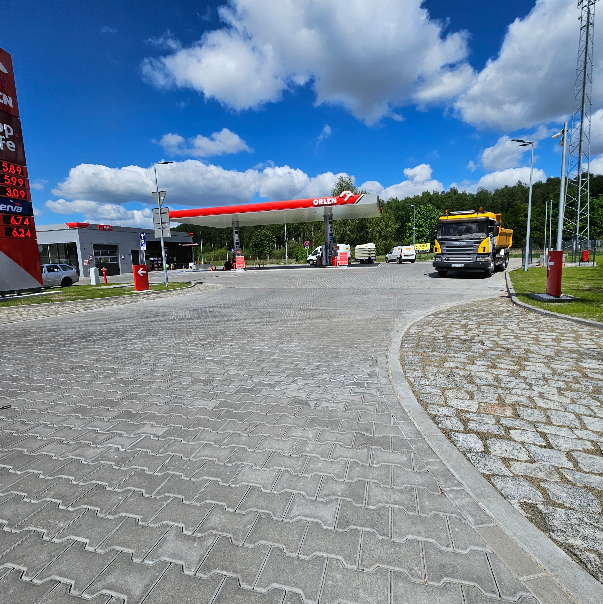Szara kostka brukowa na stacji benzynowej Orlen w słoneczny dzień. Żółta ciężarówka w tle. Widok na dystrybutory i budynek stacji. Czysta, równa nawierzchnia.
