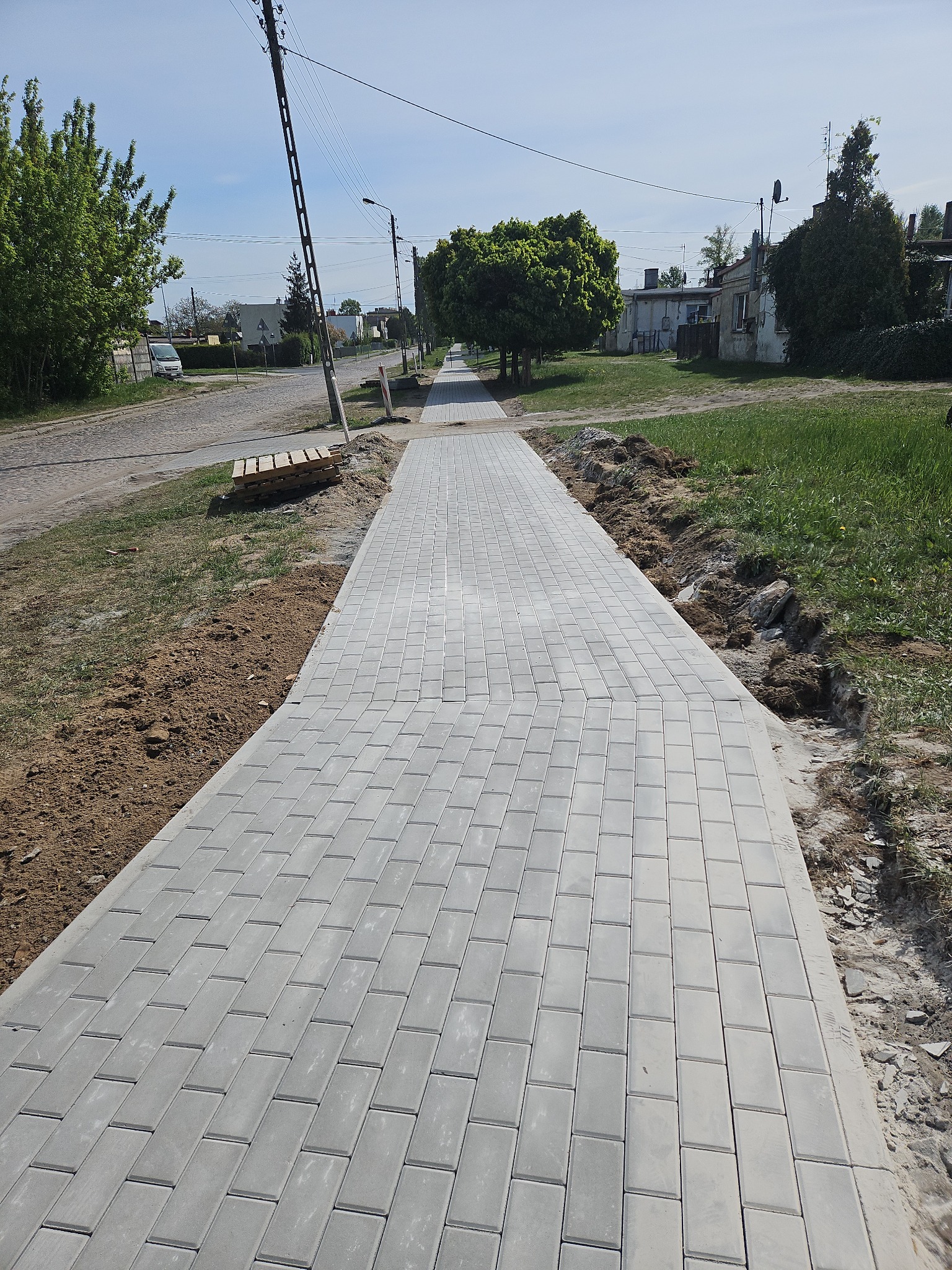 Szara kostka brukowa ułożona wzdłuż drogi, widok z perspektywy. Po bokach widoczne przygotowane podłoże i paleta. Słoneczny dzień, otoczenie wiejskie.