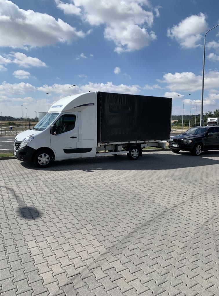 Biały bus z czarną plandeką zaparkowany na placu z kostki, obok czarny samochód osobowy. W tle widoczne latarnie i błękitne niebo z chmurami.