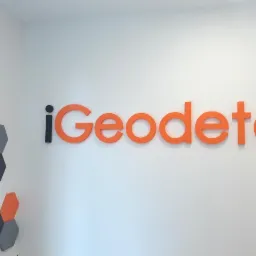Logo firmy iGeodeta w kolorze pomarańczowym, umieszczone na białej ścianie w biurze, obok dekoracji ściennej z sześciokątnych płytek w kolorach: grafitowym, pomarańczowym i białym.