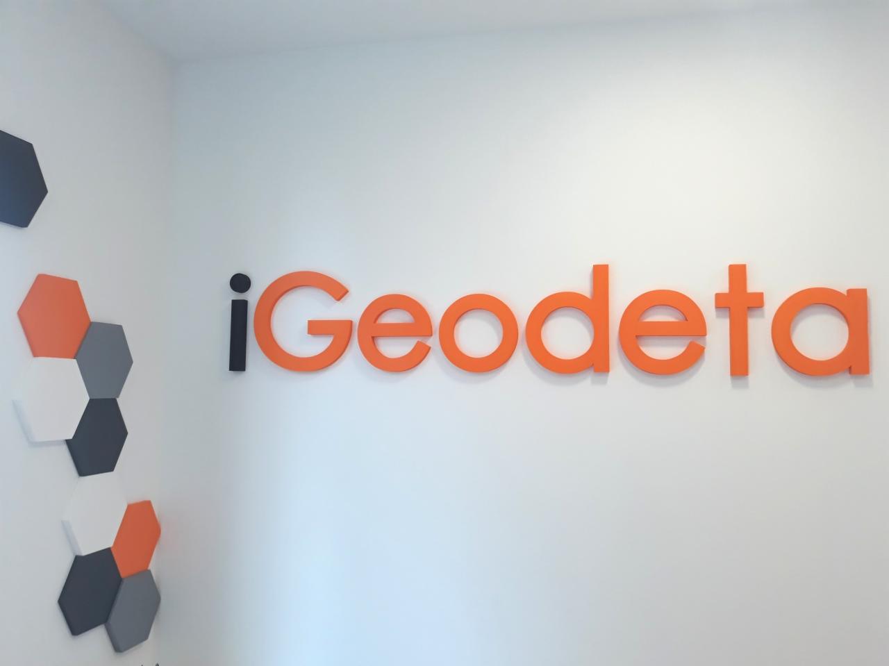 Logo firmy iGeodeta w kolorze pomarańczowym, umieszczone na białej ścianie w biurze, obok dekoracji ściennej z sześciokątnych płytek w kolorach: grafitowym, pomarańczowym i białym.