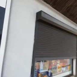 Zamknięta, grafitowa roleta zewnętrzna zabezpieczająca okno wystawowe sklepu z artykułami papierniczymi i zabawkami, widoczny fragment szyldu LOTTO.