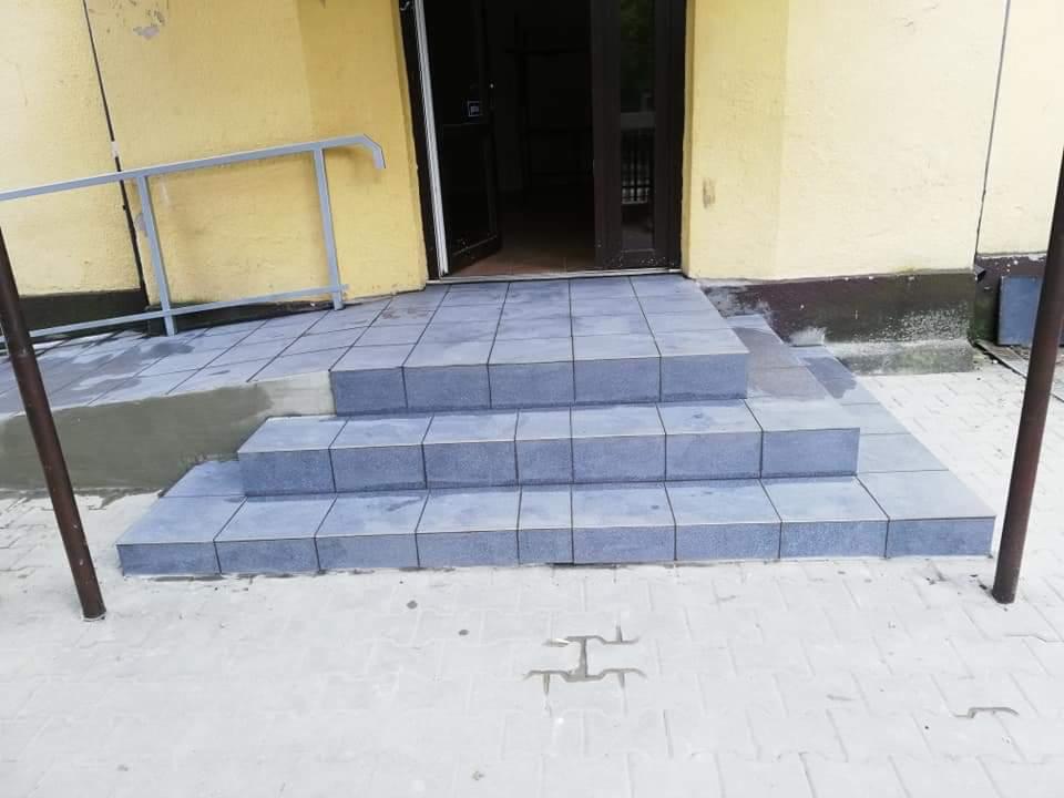 Nowe, szare płytki ceramiczne na schodach wejściowych do budynku, z rampą dla niepełnosprawnych i metalową barierką, widoczne ślady po fugowaniu.