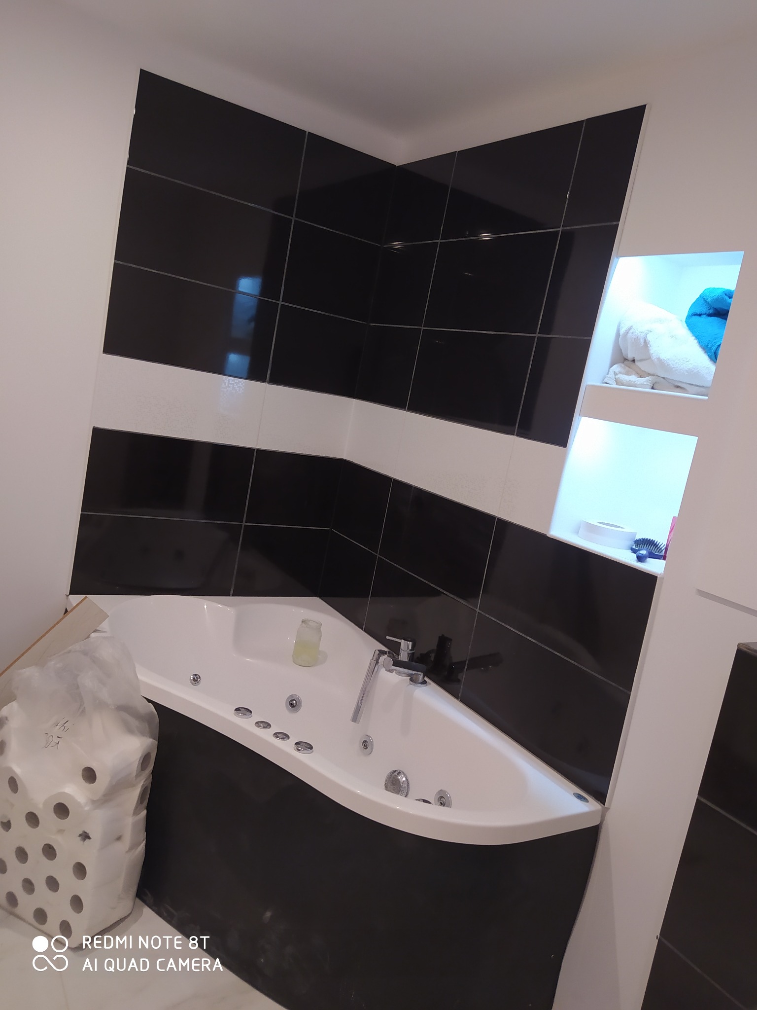 Narożna wanna z hydromasażem, obudowana czarną glazurą, z biało-czarną mozaiką na ścianach w tle, uzupełniona wnęką z półkami i akcesoriami łazienkowymi; widoczny pakiet rolek papieru toaletowego...