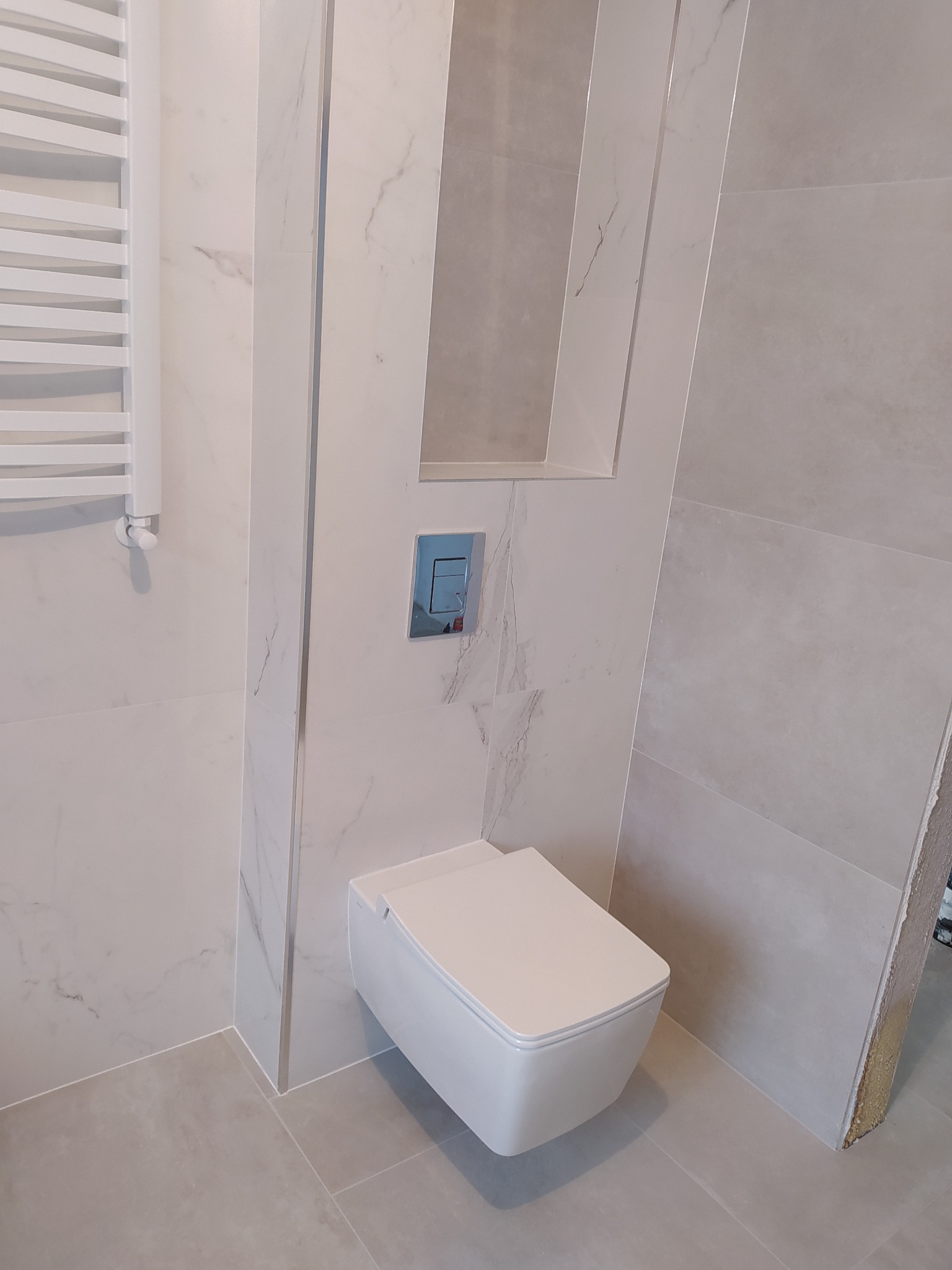 Nowoczesna toaleta podwieszana z chromowanym przyciskiem spłukującym, wbudowana w ścianę wykończoną płytkami imitującymi marmur, z wnęką dekoracyjną powyżej i grzejnikiem drabinkowym po lewej stronie.