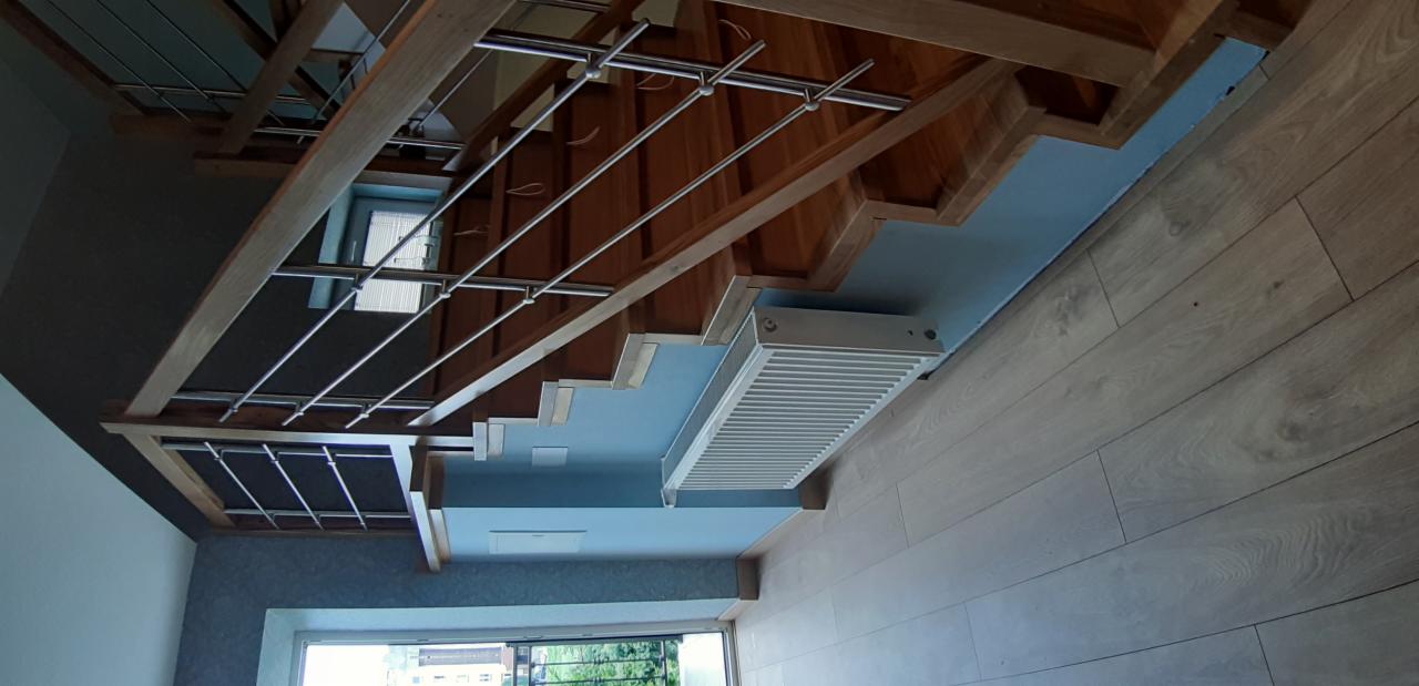 Widok od dołu na drewniane schody z metalowymi poręczami, z widocznymi elementami konstrukcyjnymi i grzejnikiem pod schodami, w jasnym wnętrzu.