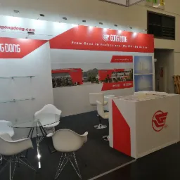 Stoisko targowe z logo firmy Gong Dong, zawierające szklany stolik otoczony białymi krzesłami, półki oraz ścianę z grafiką przedstawiającą budynek firmy i jej produkty.