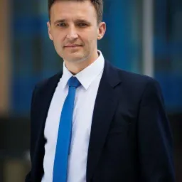 Portret mężczyzny w ciemnym garniturze, białej koszuli i niebieskim krawacie, stojącego na tle błękitnej ściany biurowca.