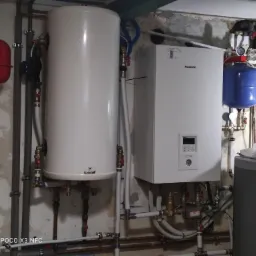 Instalacja grzewcza z białym piecem gazowym Panasonic, zbiornikiem Galmet, czerwonym i niebieskim naczyniem przeponowym, widoczne rury miedziane i plastikowe, zamontowana na surowej ścianie...