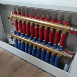 Rozdzielacz ogrzewania podłogowego w szafce, z czerwonymi i niebieskimi rurami oraz mosiężnymi elementami, widoczne przepływomierze na górnej belce.