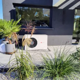 Biała jednostka zewnętrzna urządzenia grzewczego z widocznym logo Saunier Duval, umieszczona na betonowym patio w otoczeniu dekoracyjnych traw i roślin doniczkowych, z nowoczesną architekturą domu...