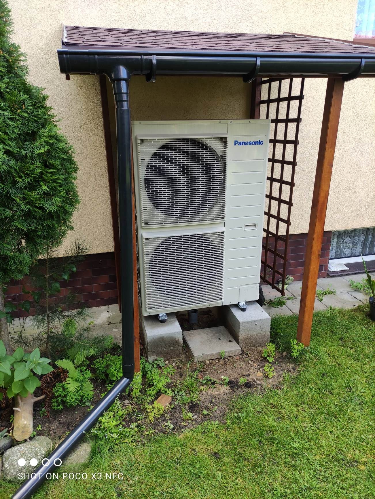 Zewnętrzna jednostka pompy ciepła Panasonic, umieszczona na betonowych bloczkach pod zadaszeniem, obok rynny spustowej i drewnianej konstrukcji kratownicy w ogrodzie.