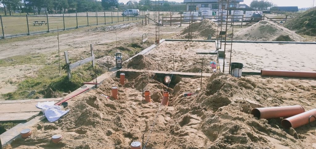 Fundament domu w trakcie budowy, widoczne pionowe rury kanalizacyjne wystające z piasku, przygotowane pod wylewkę, zarys fundamentów, w tle teren budowy i budynki.