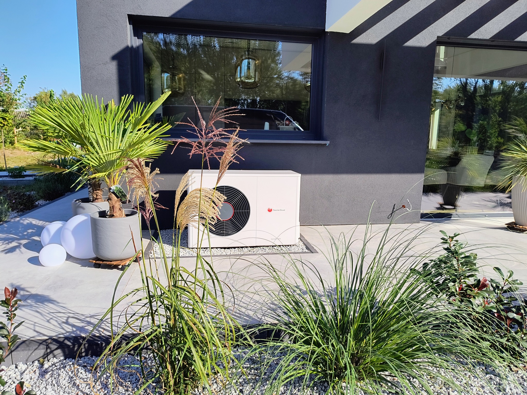 Biała jednostka zewnętrzna urządzenia grzewczego z widocznym logo Saunier Duval, umieszczona na betonowym patio w otoczeniu dekoracyjnych traw i roślin doniczkowych, z nowoczesną architekturą domu...