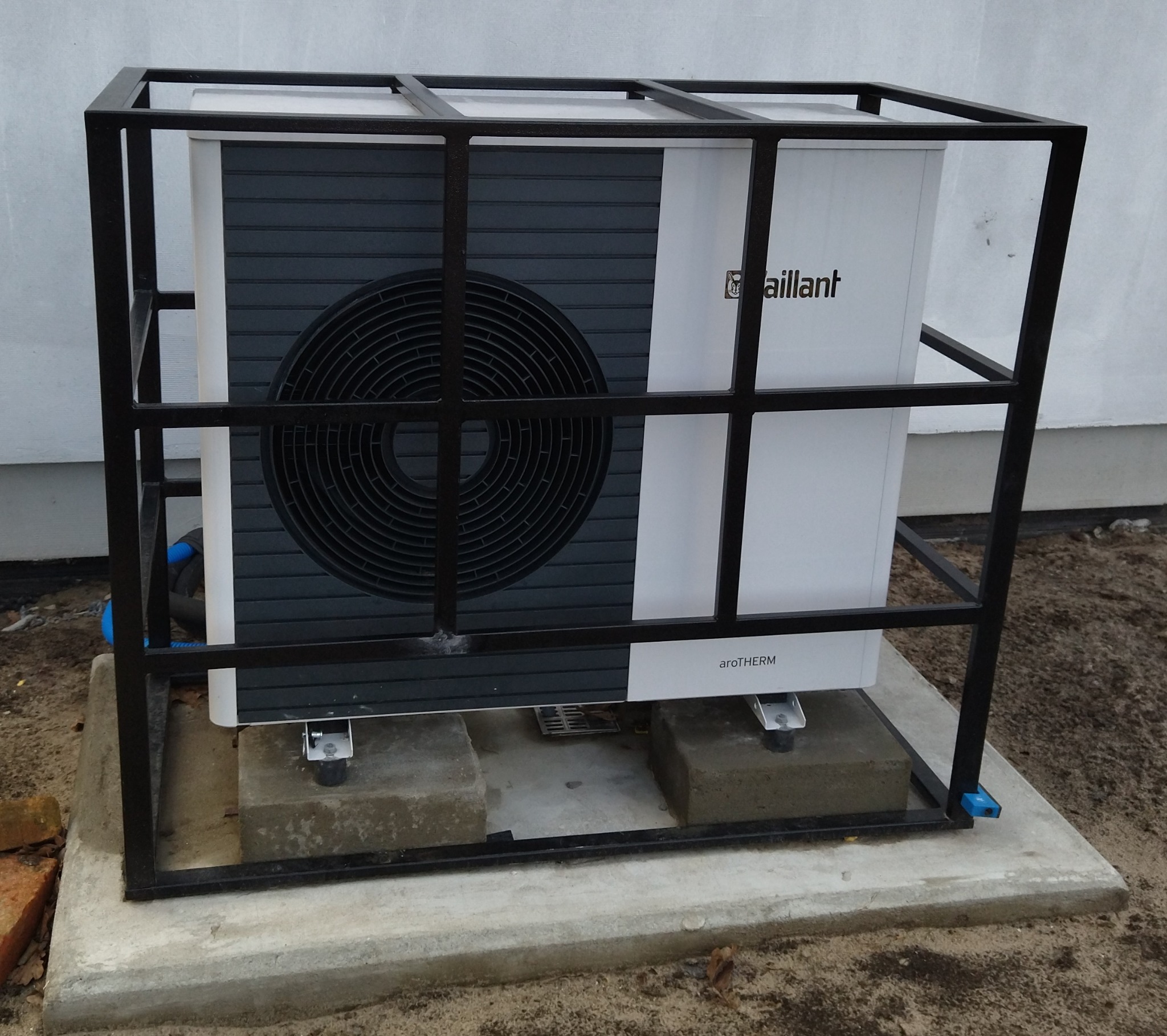 Biała pompa ciepła Vaillant aroTHERM, umieszczona w czarnej metalowej klatce ochronnej, stoi na betonowym fundamencie i kostkach, widok z boku.