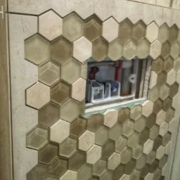 Wykończenie ściany heksagonalnymi płytkami ceramicznymi w odcieniach beżu i brązu, z widocznym otworem rewizyjnym i instalacją hydrauliczną w tle.