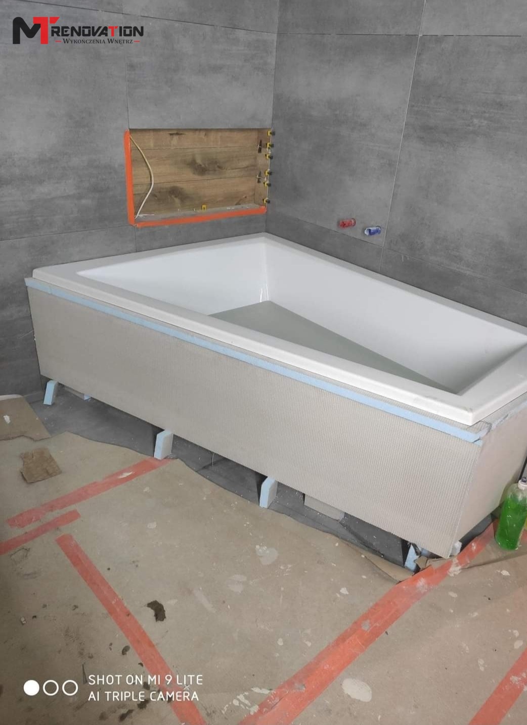 Biała wanna obudowana płytami przygotowana do wykończenia, widoczne szare płytki na ścianie, fragment instalacji wodnej i logo firmy na górze zdjęcia.