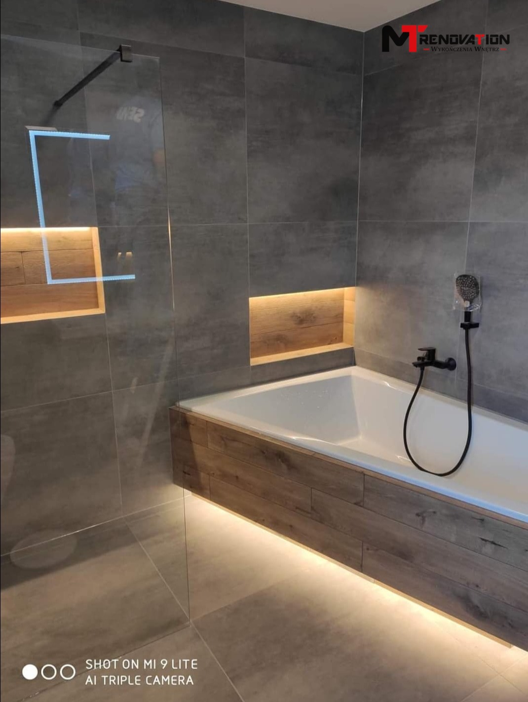 Nowoczesna łazienka z szarymi płytkami, podświetlaną wanną obudowaną drewnem i szklaną kabiną prysznicową, z logo firmy w rogu.