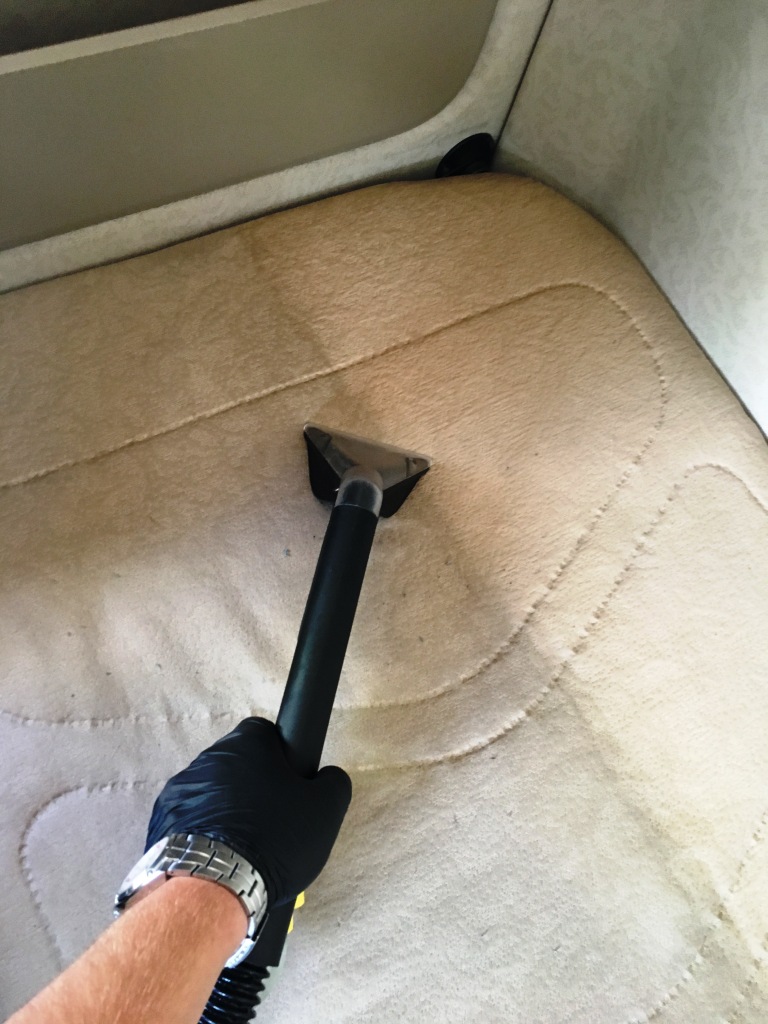 Czyszczenie materaca w kamperze za pomocą odkurzacza piorącego, widoczna ręka w czarnej rękawicy i srebrny zegarek na nadgarstku.