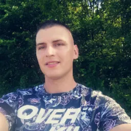 Młody mężczyzna z piercingiem brwi uśmiecha się na tle zieleni. Ubrany w koszulkę z nadrukiem czaszek. Portret w naturalnym oświetleniu.