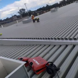 Dach z blachy trapezowej, dwaj pracownicy w odblaskowych kamizelkach przy świetliku, skrzynka narzędziowa Hilti i zwinięta miara na pierwszym planie.