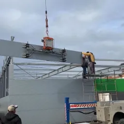 Montaż szarej płyty warstwowej za pomocą przyssawki podwieszonej do dźwigu; pracownicy na rusztowaniu korygują położenie płyty na tle konstrukcji stalowej budynku.