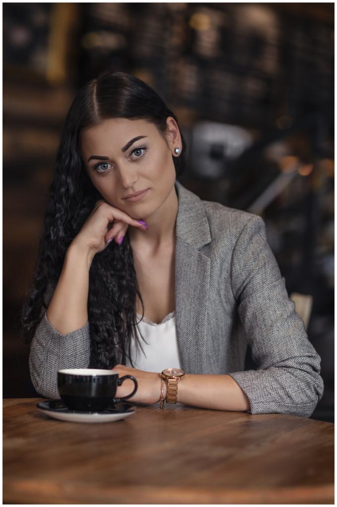 Portret kobiety w szarej marynarce, opierającej brodę na dłoni z fioletowym manicure, siedzącej przy drewnianym stole z czarną filiżanką kawy, w tle rozmyte wnętrze kawiarni.