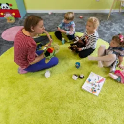 Nauczycielka siedzi na okrągłym, jasnozielonym dywanie z trójką dzieci, używając kolorowej piłki do zabawy; w tle kolorowe szafki i logo szkoły językowej.