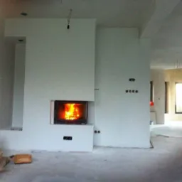 Nowoczesny kominek z paleniskiem w obudowie z jasnego tynku w surowym wnętrzu, widoczne przygotowane drewno do palenia.