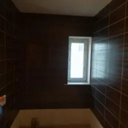 Brązowe, prostokątne płytki ułożone na ścianach w łazience, widoczne białe okno i przygotowane podłoże pod wannę lub prysznic.