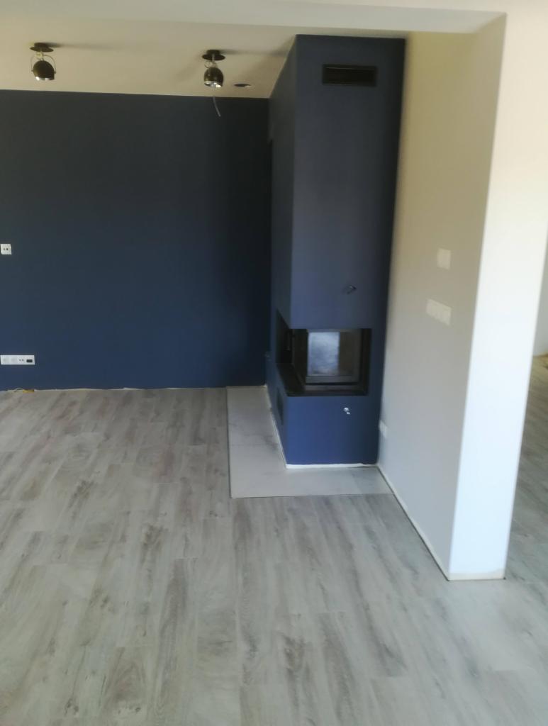 Nowoczesny, minimalistyczny kominek w kolorze granatowym, wbudowany w ścianę, z jasną podłogą imitującą drewno i ciemnogranatową ścianą w tle.