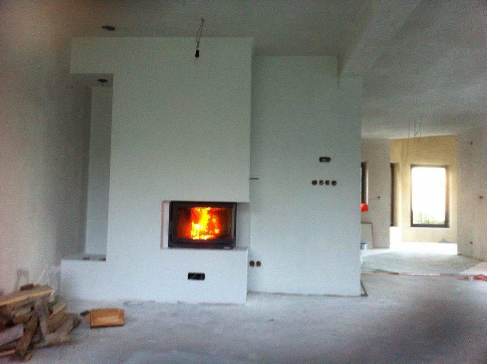 Nowoczesny kominek z paleniskiem w obudowie z jasnego tynku w surowym wnętrzu, widoczne przygotowane drewno do palenia.